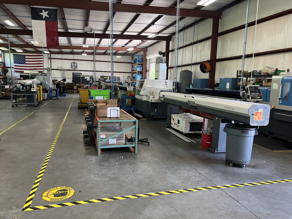 TXSwiss precision Swiss CNC shop floor in Conroe, Texas with bar-fed lathes, racks of bar stock, and US and Texas flags overhead